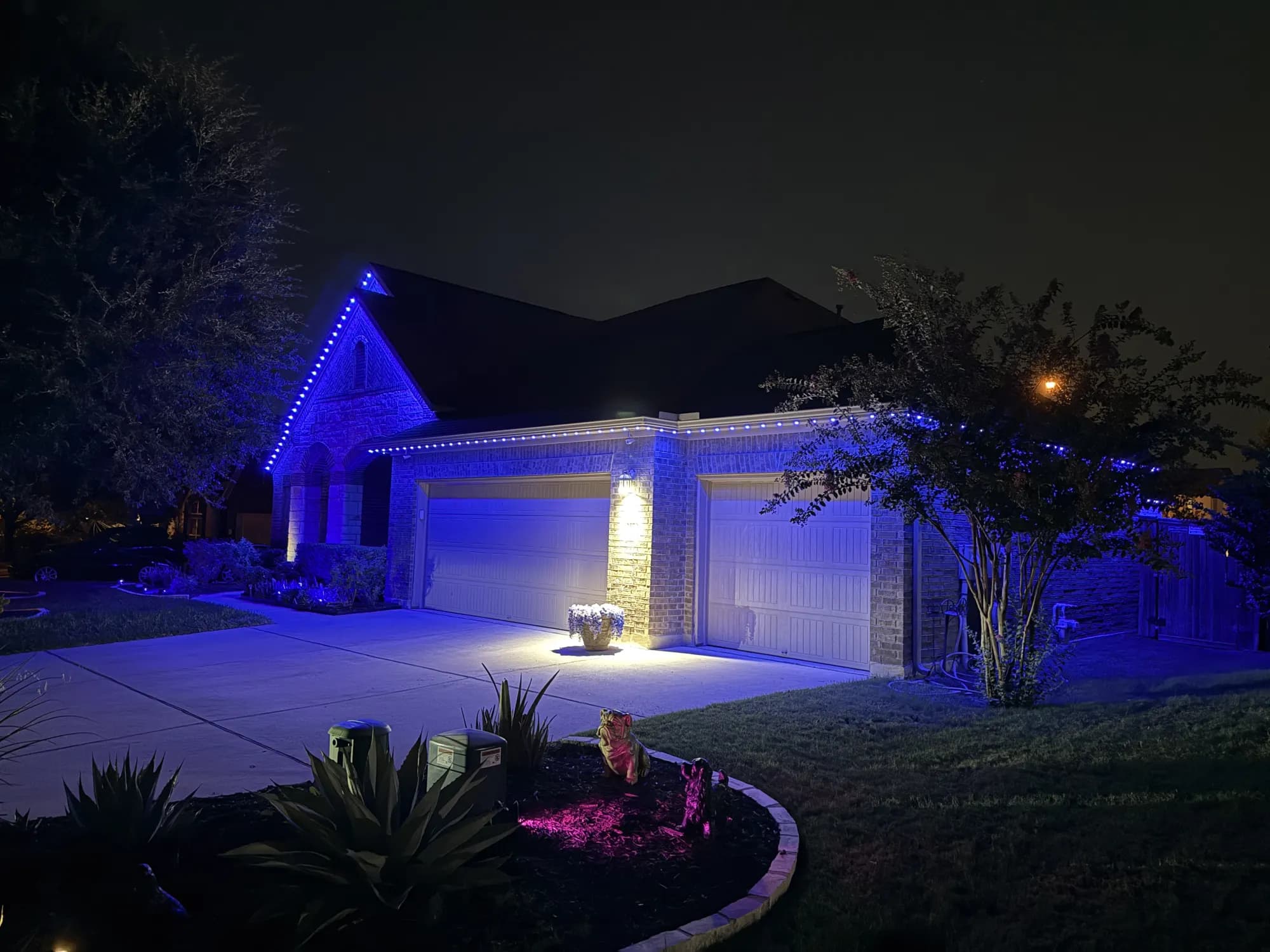 Blue TruLight lighting along the gable of a brick home at night, with landscape lights in the front bed.