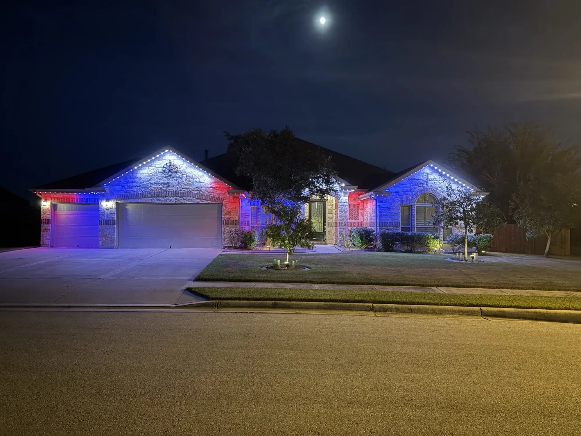 Red, white and blue TruLight roofline lighting across a single-story ranch home at dusk under a full moon.