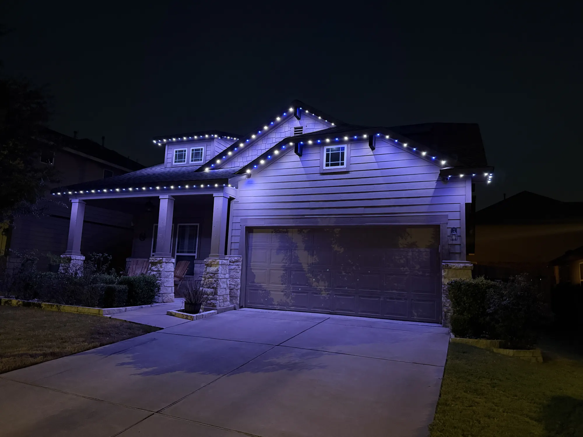 Blue and white TruLight roofline lighting on a home with siding and stone porch columns at night.