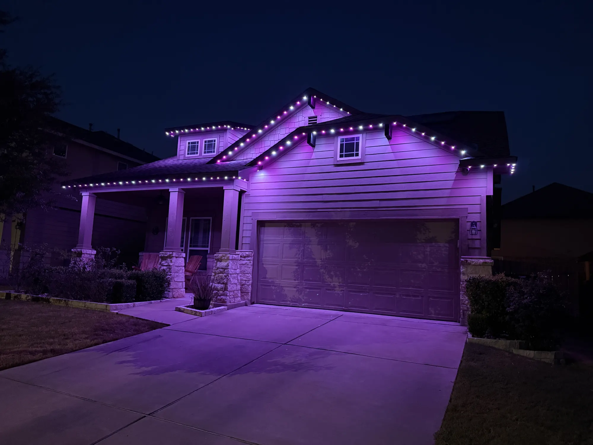 Purple and white TruLight roofline lighting on a home with siding and stone porch columns at night.