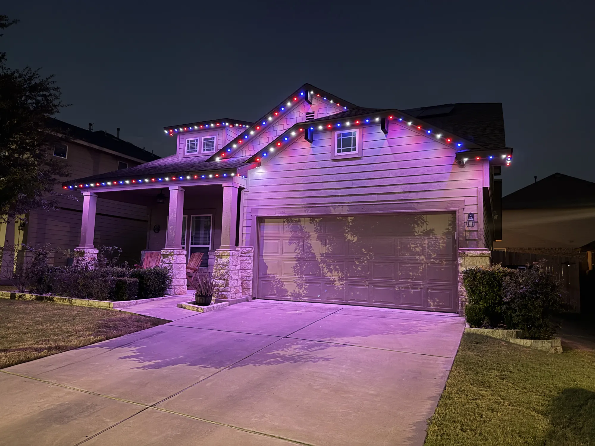 Red, white and blue TruLight roofline lighting on a home with siding and stone porch columns, with a purple glow on the siding.