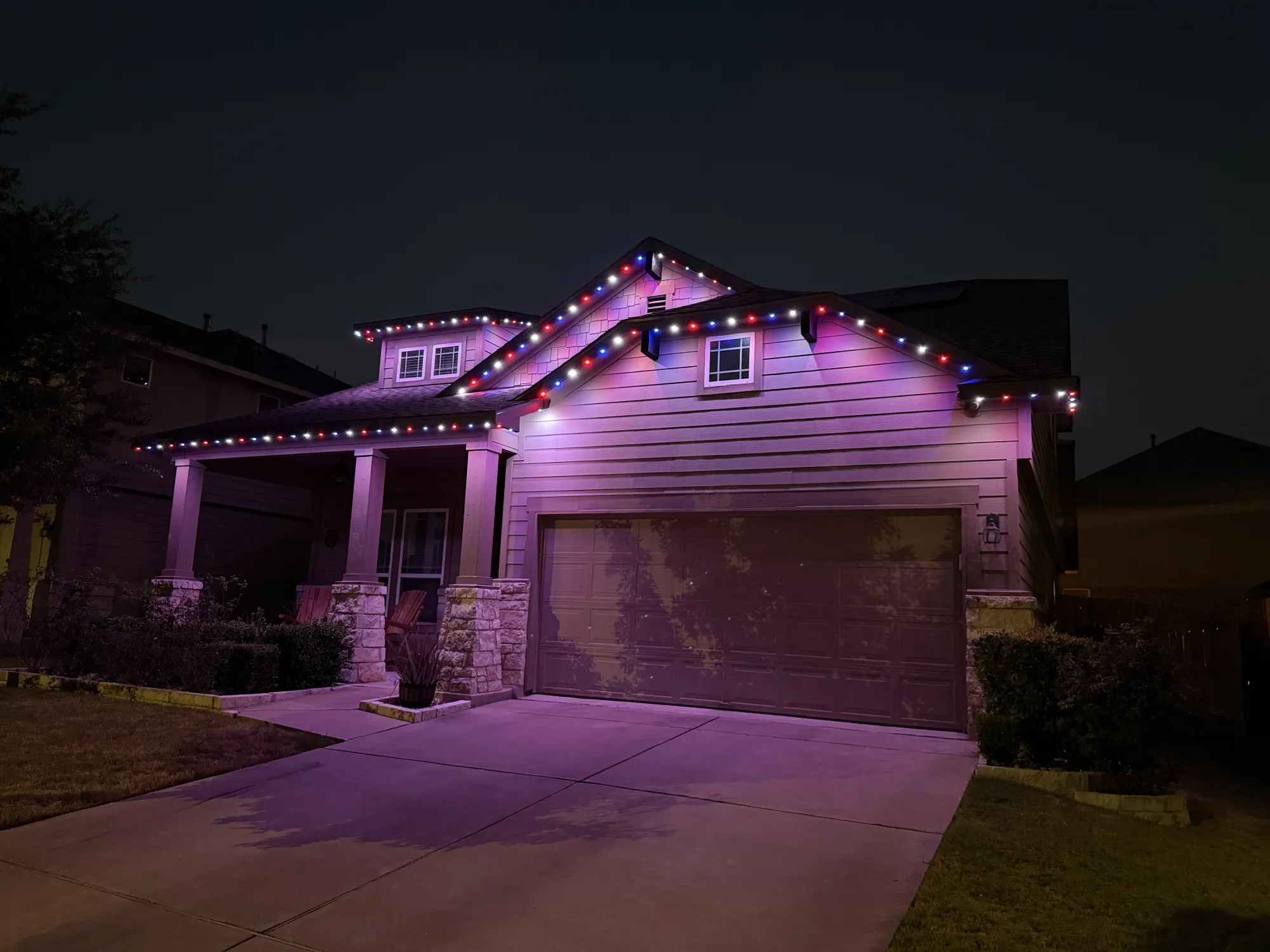 Red, white and blue TruLight roofline lighting on a home with siding and stone porch columns at night.