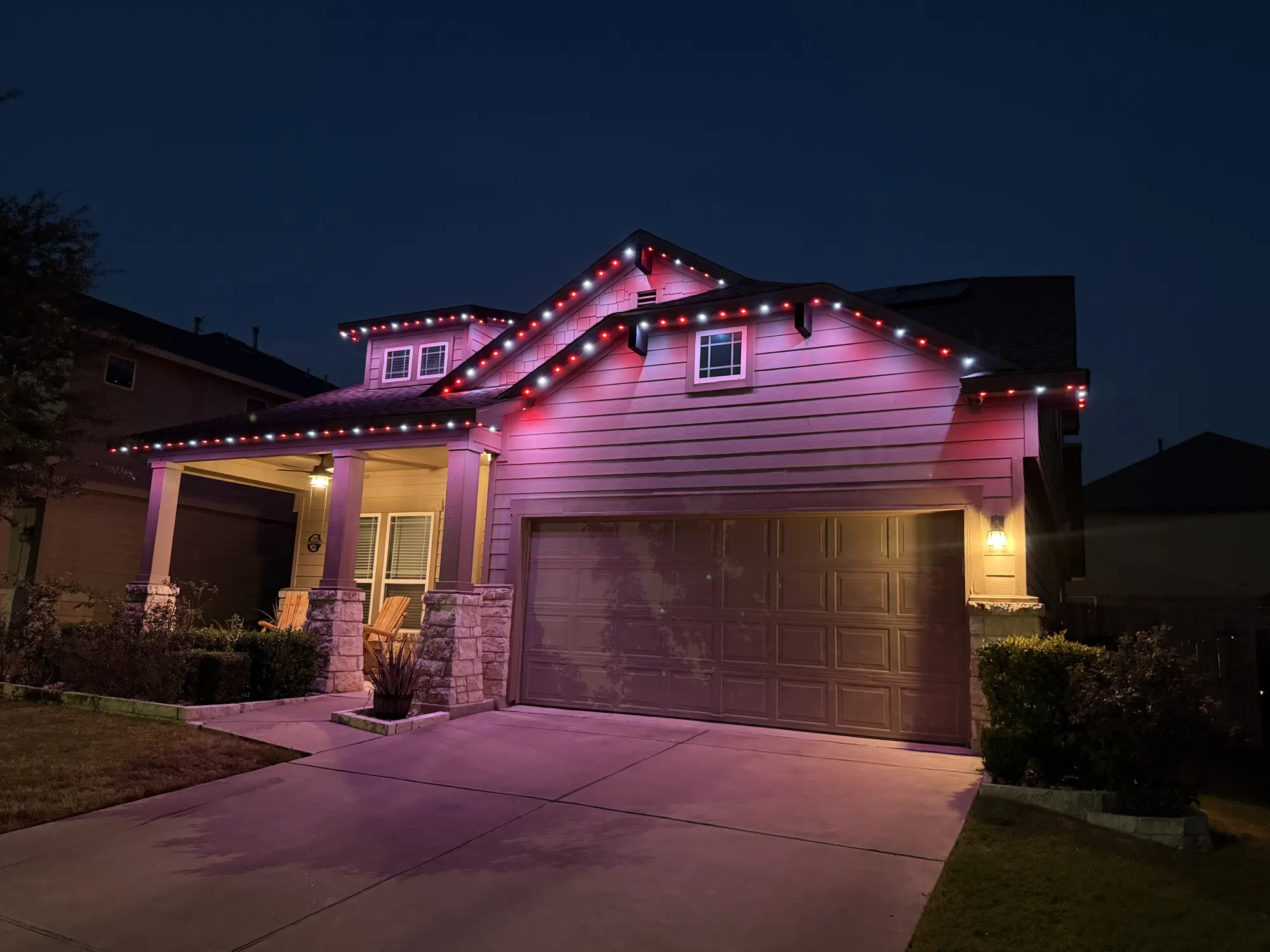 Red and white TruLight roofline lighting with a pink glow on a home with siding and stone porch columns at night.
