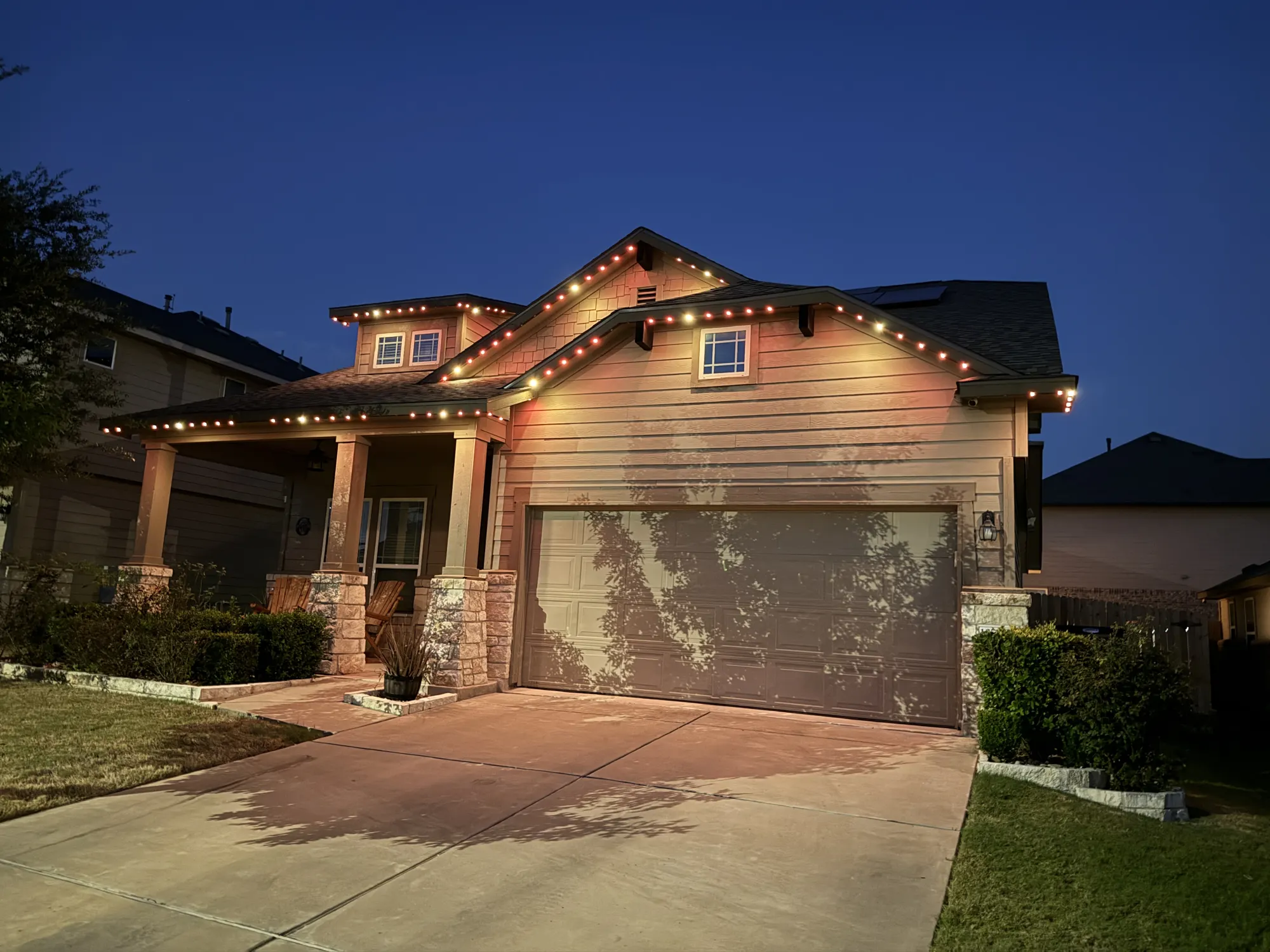 Warm-white TruLight roofline lighting on a home with siding and stone porch columns under a deep blue dusk sky.