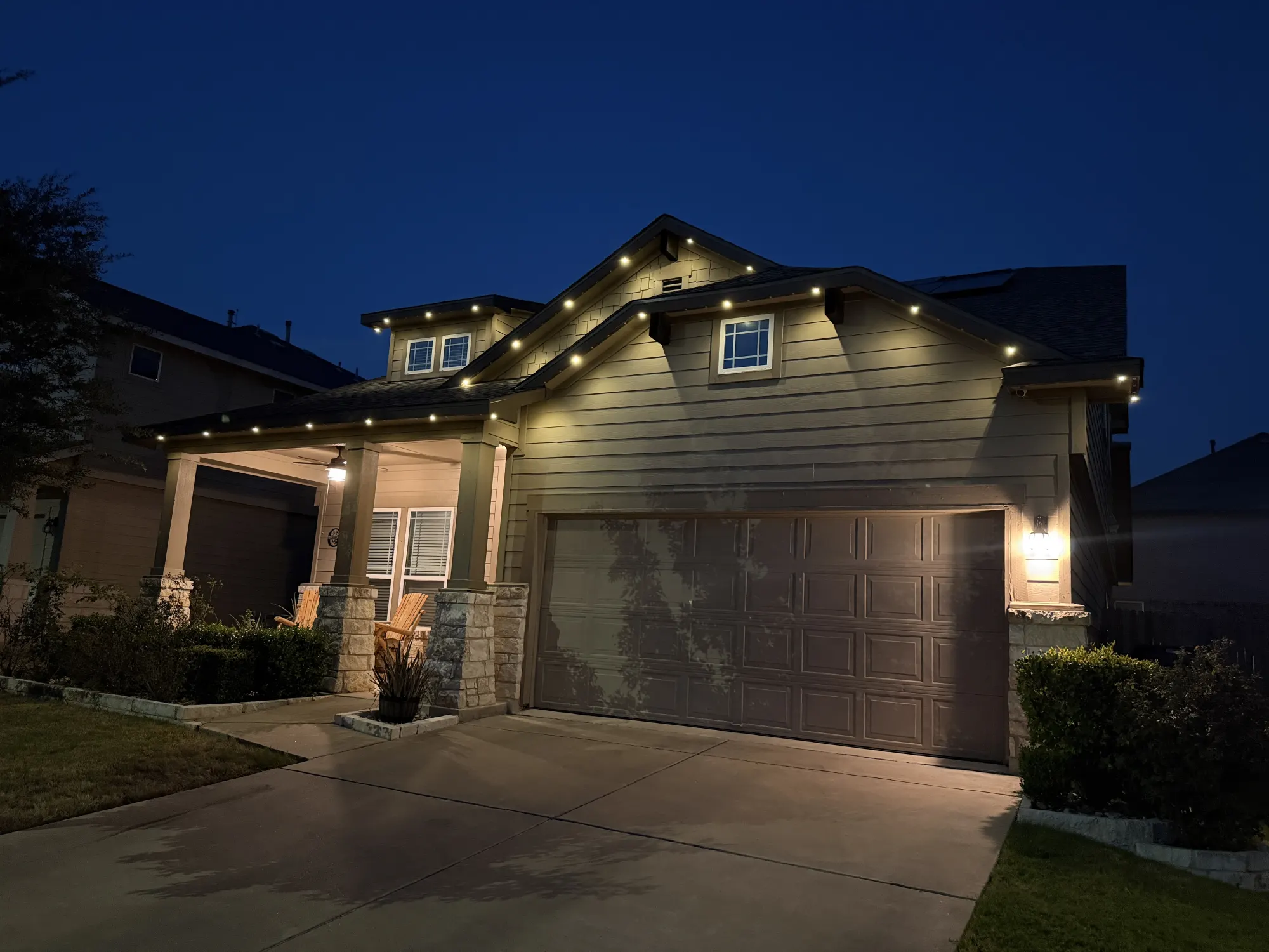 Warm-white TruLight roofline lighting on a home with siding and stone porch columns at dusk.